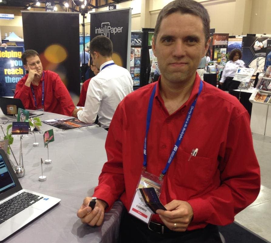 Andrew DePaula, pictured in the ASI exhibition hall, holding a regular USB stick in one hand and a paper USB card in the other. (Andrew McChesney / AR)