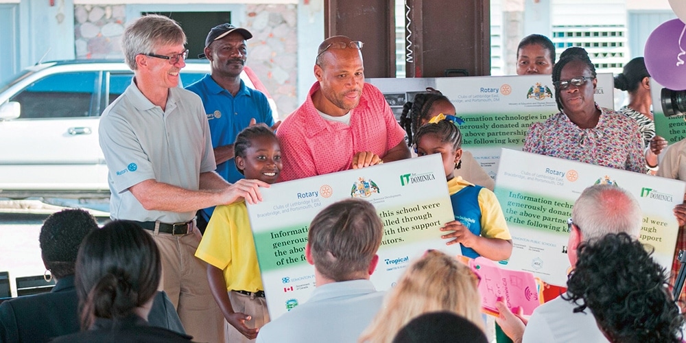 Maurice Hollingsworth of Lethbridge, Alberta, Canada (far left) attends the handover ceremony celebrating the installation of learning technologies in Dominica in November 2019. The Rotary Club of Portsmouth, Dominica had this sign made to show all of the partners, including A Better World Canada, that helped make the program a success. [Photo: A Better World Canada]