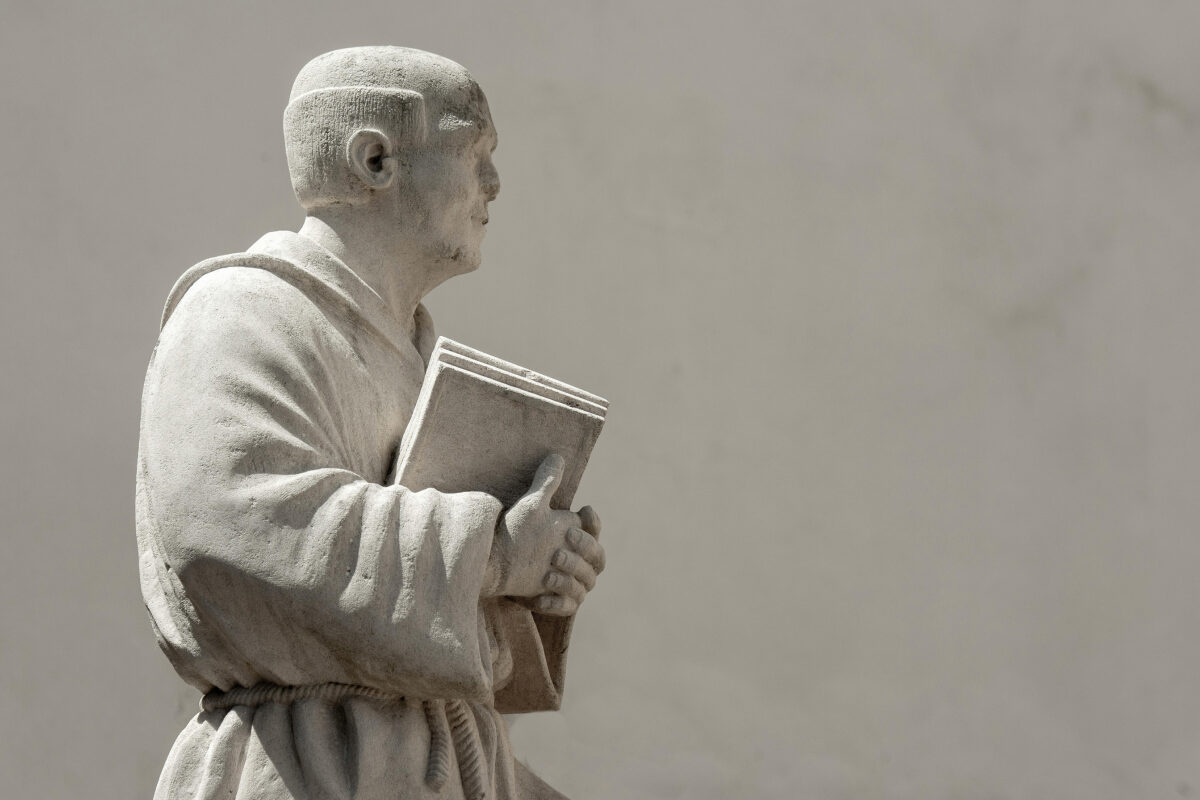 Stone statue of a monk holding a closed book against his chest, wearing a simple robe.