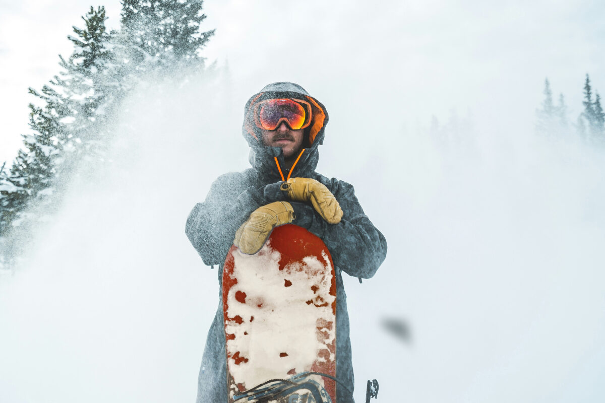 A snowboarder wearing goggles and winter gear stands in blowing snow, holding a snow-covered red snowboard upright.
