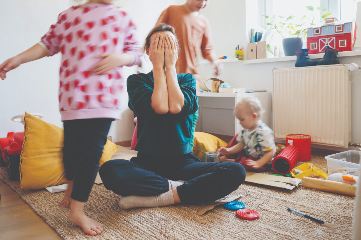 Mom sitting on the floor with hands covering her face while children play energetically around her in a messy room.