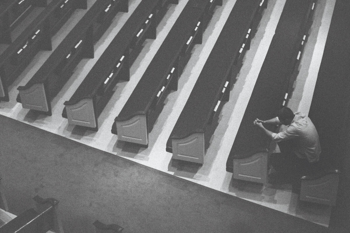 Person sitting alone in a church pew with head bowed and hands clasped in prayer.