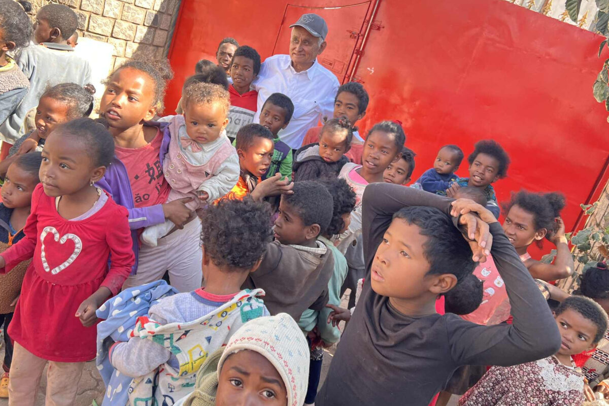 A group of curiosity and energy-filled children gather closely outside near a red gate