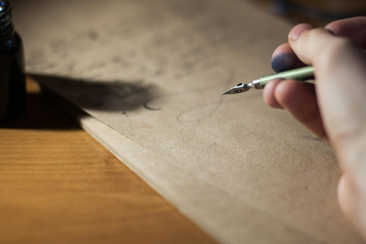 A hand holding a dip pen with ink-stained fingers, writing on brown paper beside an ink bottle.