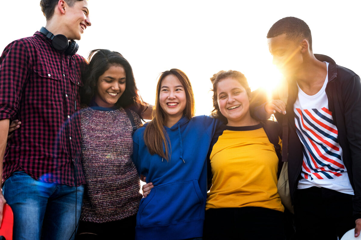 A group of smiling teenagers stands close together with their arms around each other in bright sunlight