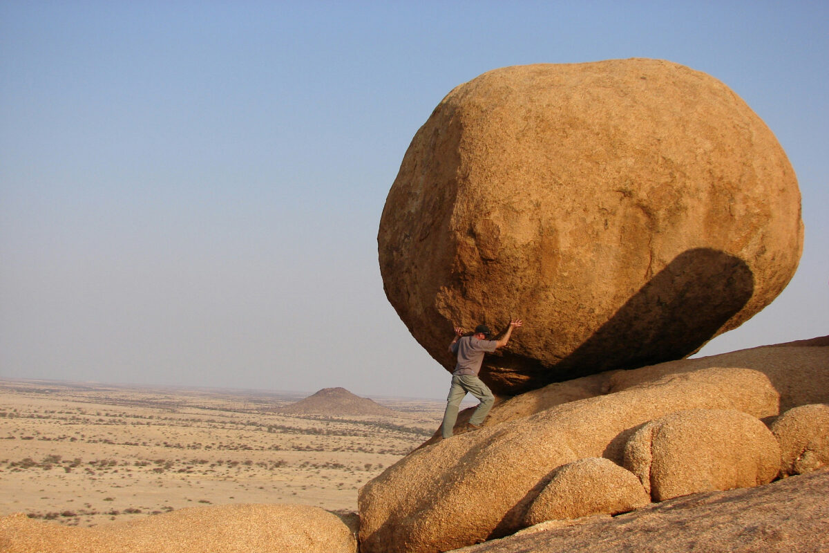 A man braces himself against a massive boulder perched on a rocky slope, as if trying to push it.