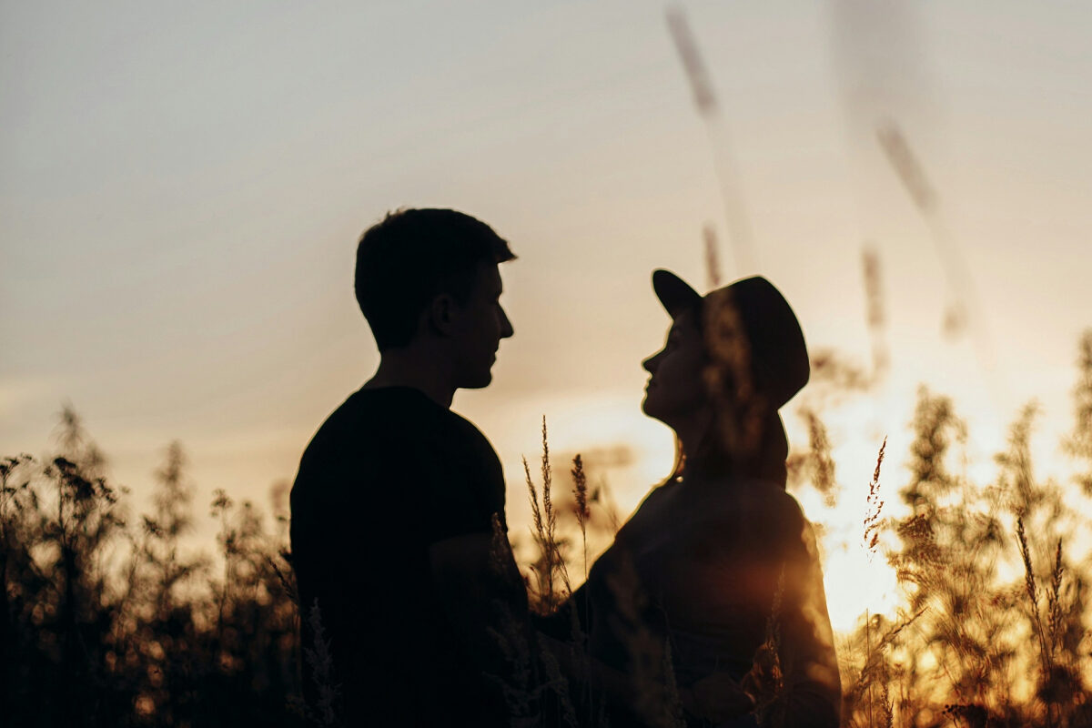 A photo of a couple standing face to face in a field at sunset, their silhouettes softly lit by the fading light.