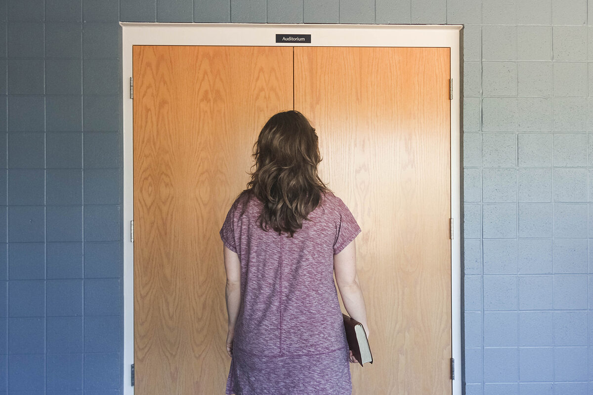 A photo of a woman standing before closed church doors holding a book, suggesting a moment of decision or entry.